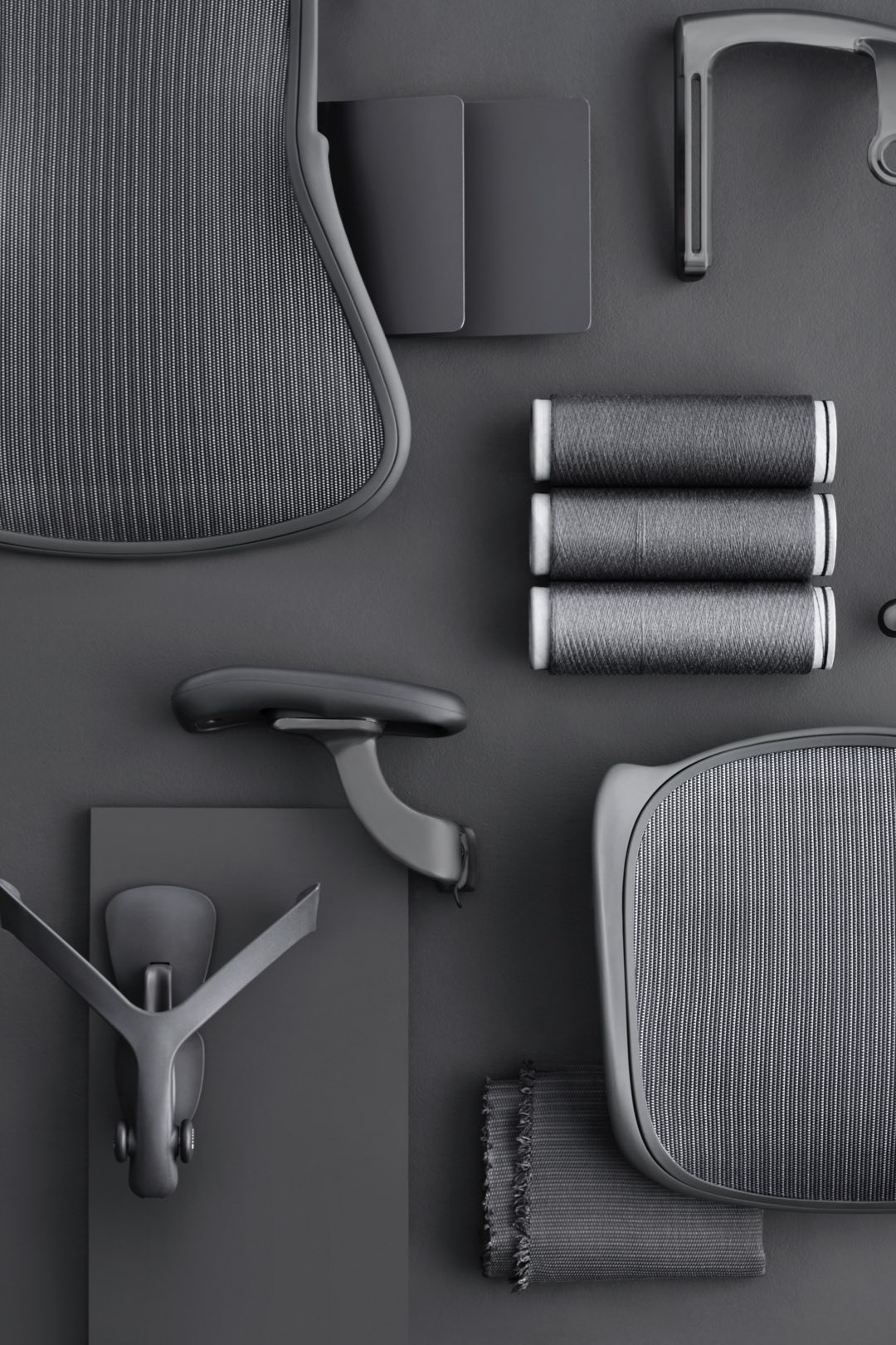 Individual parts of the new Aeron chair; including PostureFit SL pads, an armrest, set, and spools of Pellicle thread—all in Carbon finish—rest on a grey surface