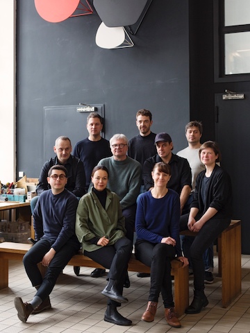 The men and women of Studio 7.5 sit grouped together on wooden benches in their workspace.