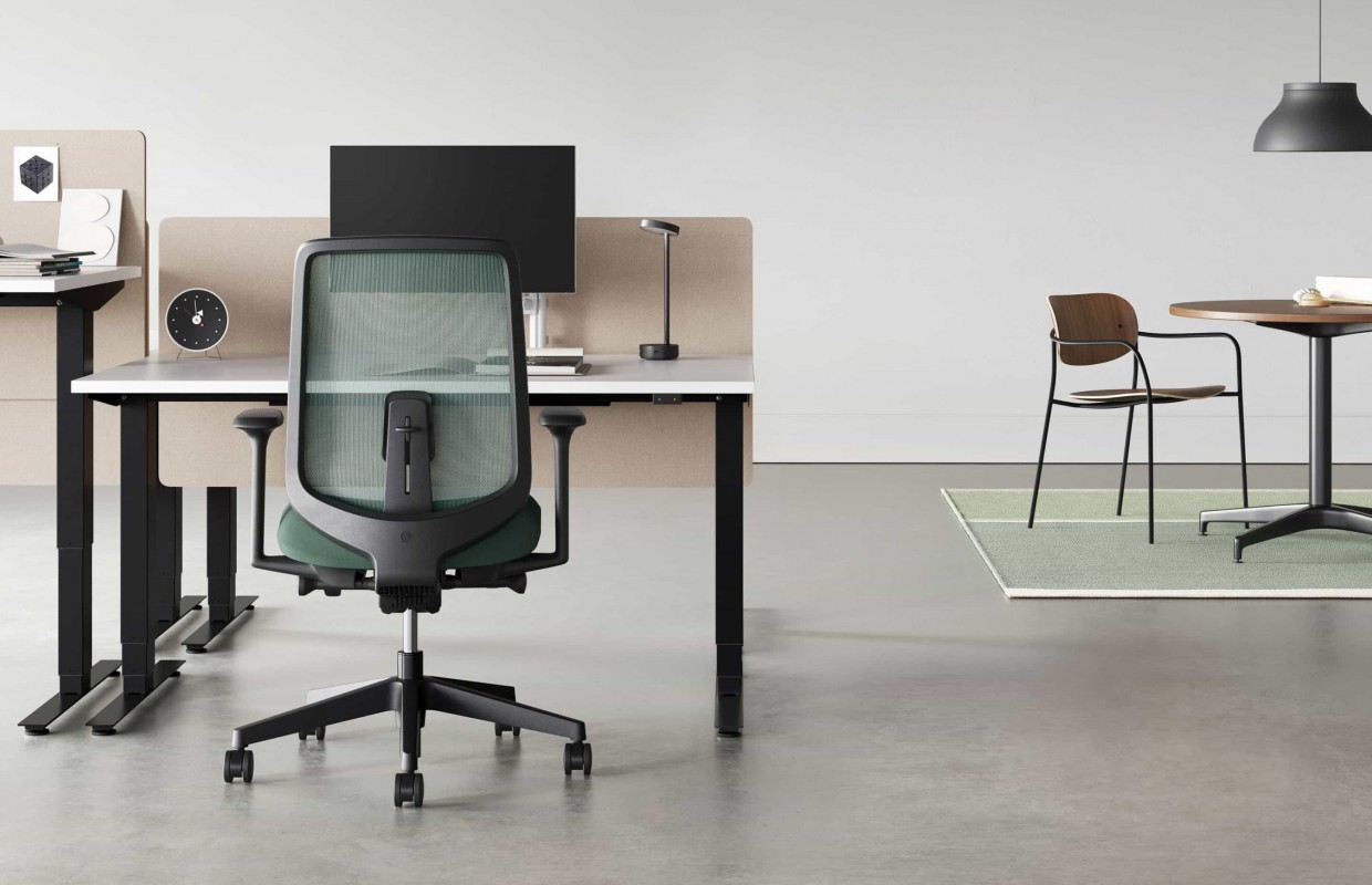 A black Verus Chair with dark green suspension back pulled up to a black Nevi Sit-Stand Desk with a Civic Table and Portrait Chair in the background.